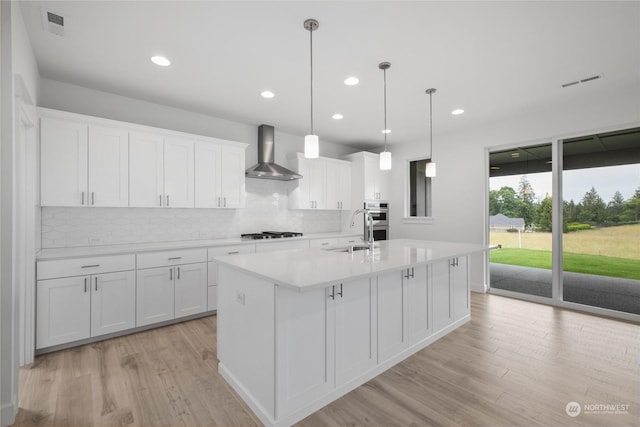 kitchen with a kitchen island with sink, hanging light fixtures, wall chimney exhaust hood, light hardwood / wood-style floors, and white cabinetry