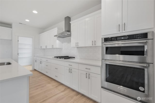 kitchen featuring white cabinets, light hardwood / wood-style flooring, wall chimney exhaust hood, light stone countertops, and stainless steel appliances