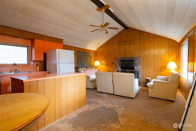 kitchen featuring ceiling fan, lofted ceiling with beams, white refrigerator, wooden walls, and a fireplace