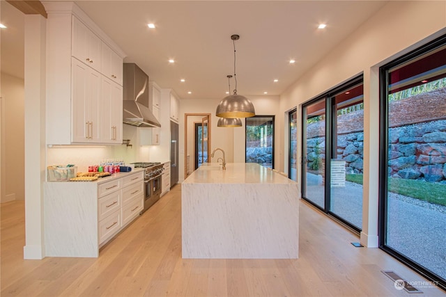 kitchen with white cabinetry, light wood-type flooring, an island with sink, pendant lighting, and wall chimney range hood