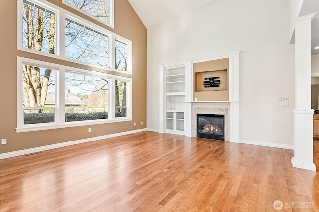 unfurnished living room featuring a high ceiling, baseboards, light wood finished floors, a tiled fireplace, and decorative columns