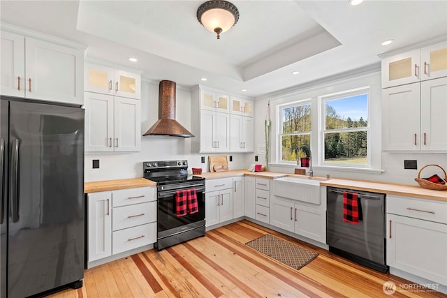 kitchen featuring light wood finished floors, stainless steel appliances, a tray ceiling, wall chimney exhaust hood, and white cabinetry