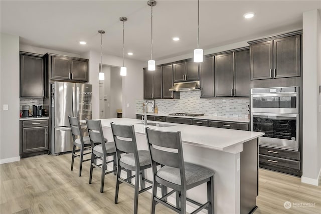 kitchen with a kitchen bar, stainless steel appliances, a kitchen island with sink, pendant lighting, and light hardwood / wood-style flooring