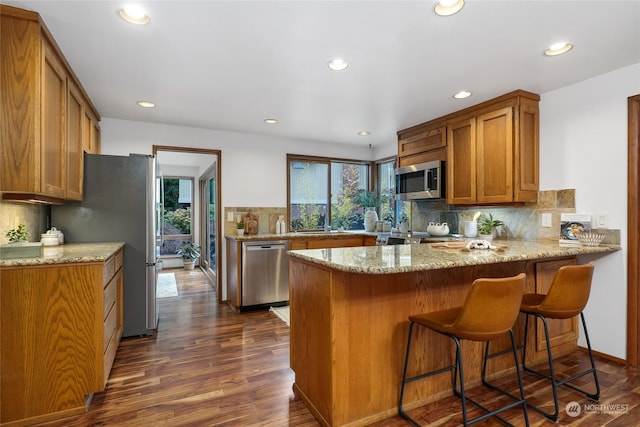 kitchen with backsplash, kitchen peninsula, light stone countertops, stainless steel appliances, and dark hardwood / wood-style flooring