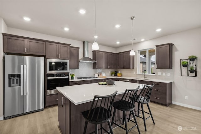 kitchen with light wood-type flooring, wall chimney exhaust hood, stainless steel appliances, decorative light fixtures, and a kitchen island