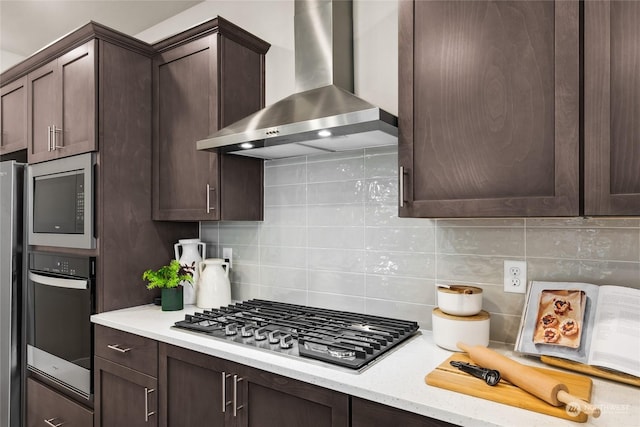 kitchen with backsplash, dark brown cabinets, stainless steel appliances, and wall chimney range hood