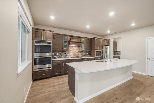 kitchen with light wood-type flooring, custom range hood, dark brown cabinetry, stainless steel appliances, and an island with sink