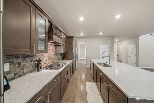 kitchen featuring a center island with sink, sink, light wood-type flooring, appliances with stainless steel finishes, and custom range hood