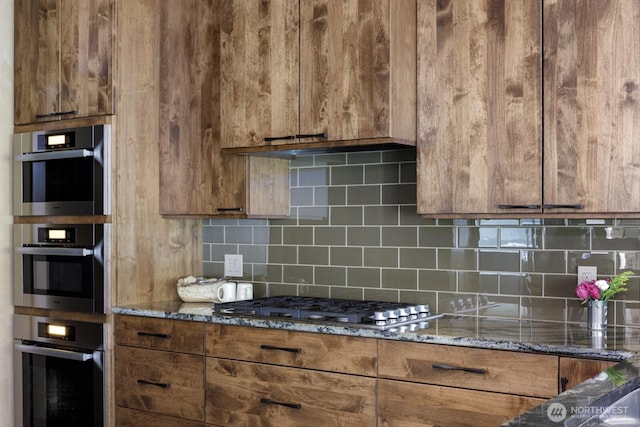 kitchen featuring brown cabinetry, dark stone countertops, stainless steel appliances, and decorative backsplash