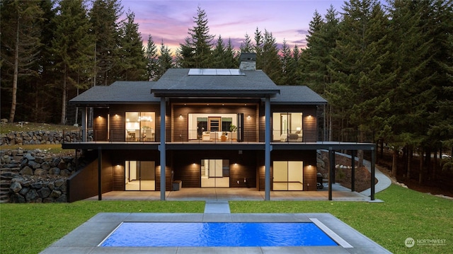 back house at dusk with solar panels, a balcony, a pool side deck, a patio area, and a lawn