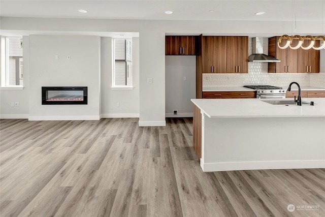 kitchen with a glass covered fireplace, light countertops, wall chimney range hood, stainless steel range with gas cooktop, and a sink