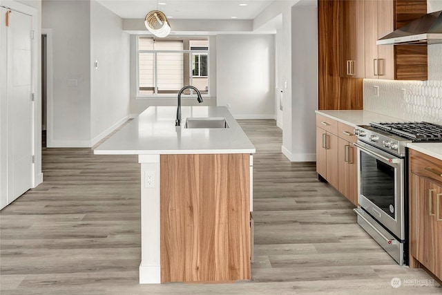 kitchen with stainless steel gas stove, an island with sink, sink, wall chimney range hood, and light wood-type flooring