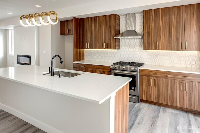 kitchen featuring wall chimney range hood, sink, stainless steel gas range, light hardwood / wood-style floors, and a center island with sink