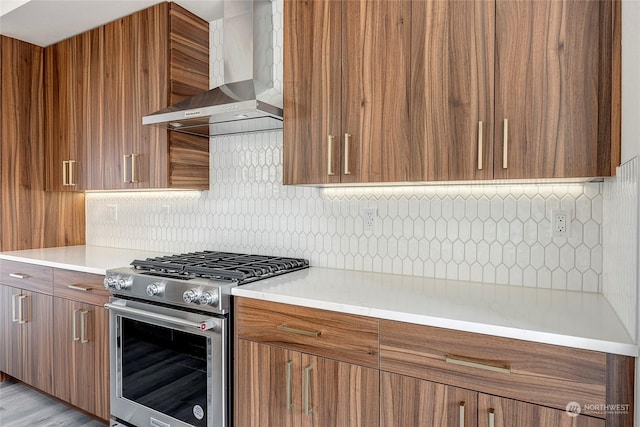 kitchen featuring brown cabinetry, stainless steel range with gas stovetop, backsplash, and wall chimney exhaust hood