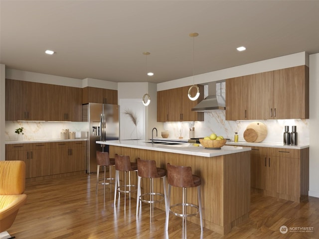 kitchen with hanging light fixtures, wood-type flooring, a center island with sink, stainless steel fridge with ice dispenser, and wall chimney exhaust hood