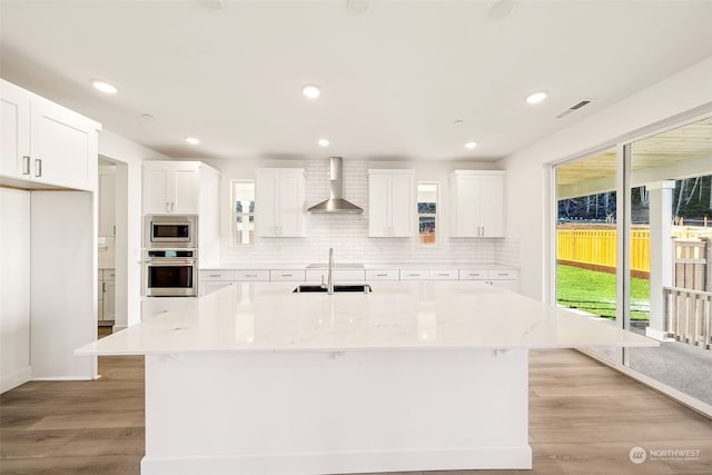 kitchen featuring wall chimney exhaust hood, stainless steel appliances, a kitchen island with sink, and white cabinets