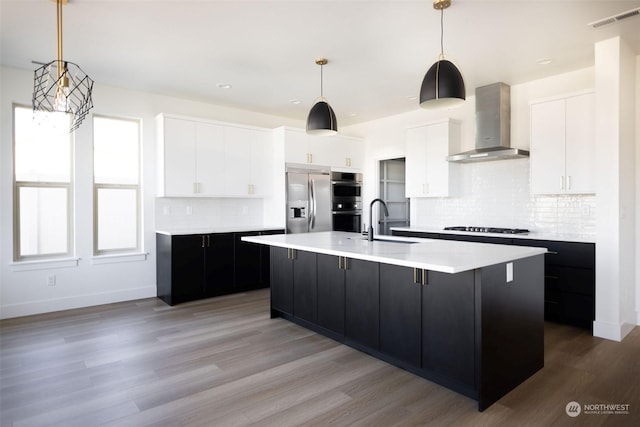 kitchen with pendant lighting, a center island with sink, stainless steel appliances, white cabinets, and wall chimney exhaust hood