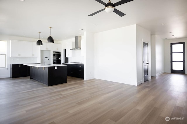 kitchen featuring decorative light fixtures, a kitchen island with sink, wall chimney exhaust hood, white cabinets, and sink