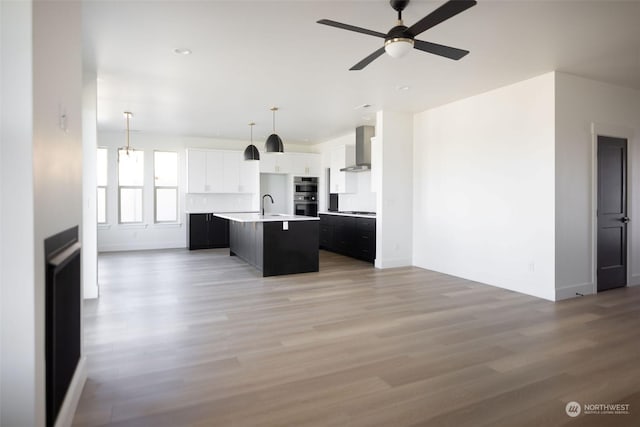 kitchen with wall chimney exhaust hood, decorative light fixtures, white cabinetry, an island with sink, and ceiling fan