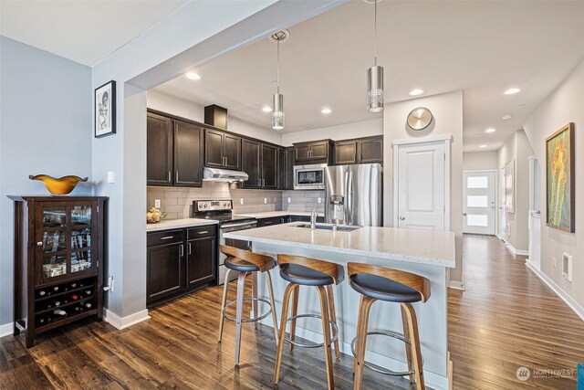 kitchen with hanging light fixtures, dark wood-type flooring, stainless steel appliances, an island with sink, and decorative backsplash