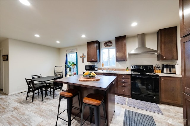 kitchen featuring dark brown cabinets, wall chimney range hood, black electric range, a center island, and light hardwood / wood-style floors