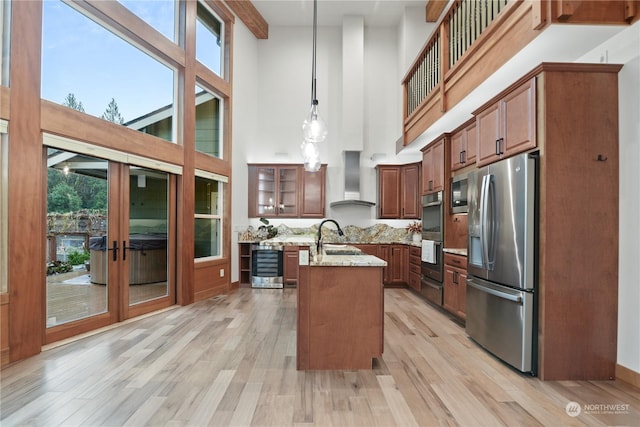 kitchen with a towering ceiling, decorative light fixtures, stainless steel appliances, a center island with sink, and wall chimney exhaust hood