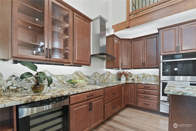 kitchen featuring wall chimney exhaust hood, light stone counters, beverage cooler, stainless steel double oven, and light wood-type flooring