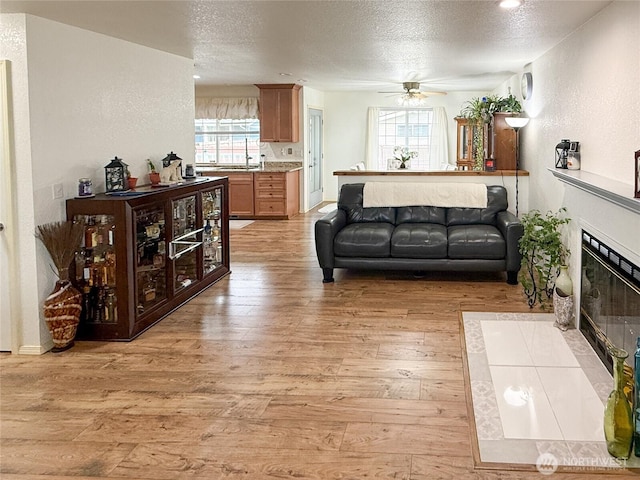 living area featuring light wood finished floors, a textured wall, a fireplace with flush hearth, ceiling fan, and a textured ceiling