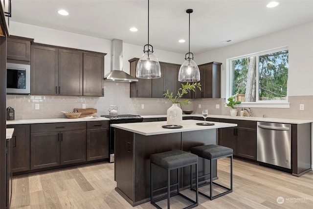 kitchen featuring wall chimney exhaust hood, hanging light fixtures, stainless steel appliances, an island with sink, and light hardwood / wood-style floors