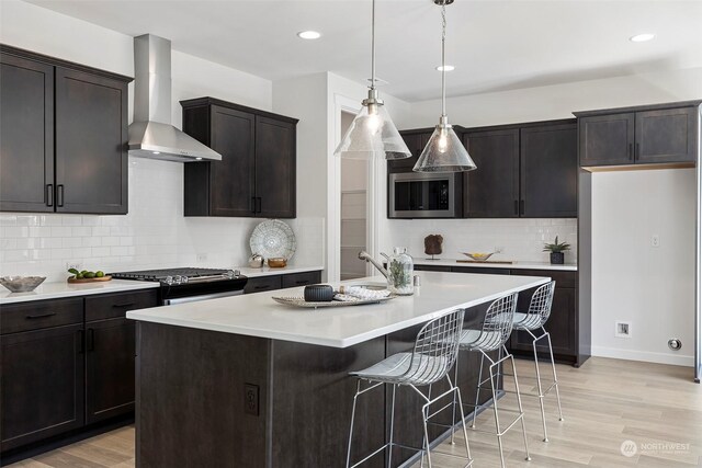 kitchen featuring wall chimney exhaust hood, stainless steel appliances, pendant lighting, a center island with sink, and light hardwood / wood-style floors