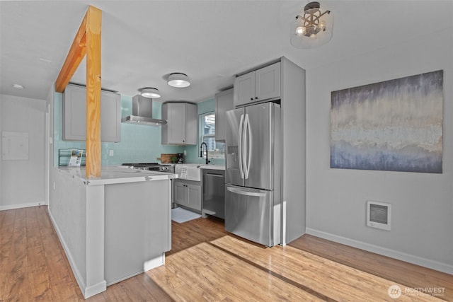 kitchen featuring wall chimney exhaust hood, appliances with stainless steel finishes, light countertops, gray cabinetry, and light wood-type flooring
