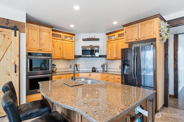 kitchen featuring sink, stainless steel appliances, a barn door, light stone counters, and a kitchen island with sink