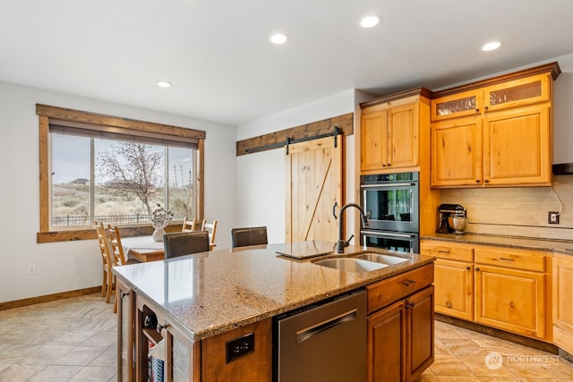 kitchen featuring appliances with stainless steel finishes, light stone counters, sink, a barn door, and an island with sink