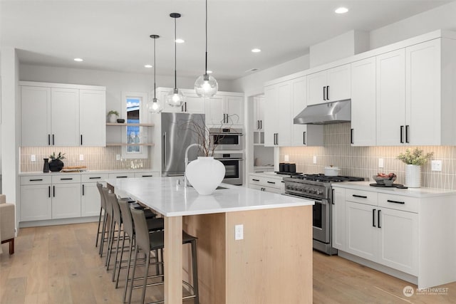 kitchen featuring under cabinet range hood, stainless steel appliances, a breakfast bar, open shelves, and a center island with sink