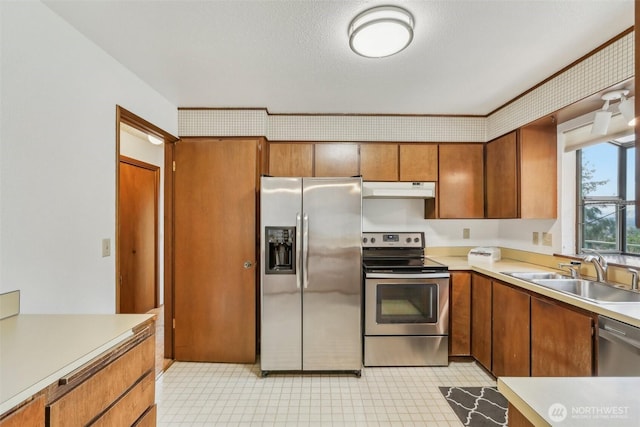 kitchen featuring a sink, light countertops, under cabinet range hood, appliances with stainless steel finishes, and brown cabinets