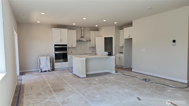 kitchen featuring recessed lighting, stainless steel double oven, a center island with sink, and range hood