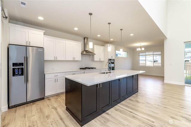 kitchen with a kitchen island with sink, white cabinets, wall chimney exhaust hood, decorative light fixtures, and stainless steel appliances