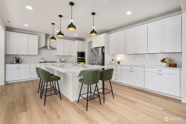 kitchen featuring pendant lighting, wall chimney range hood, light hardwood / wood-style flooring, appliances with stainless steel finishes, and white cabinetry