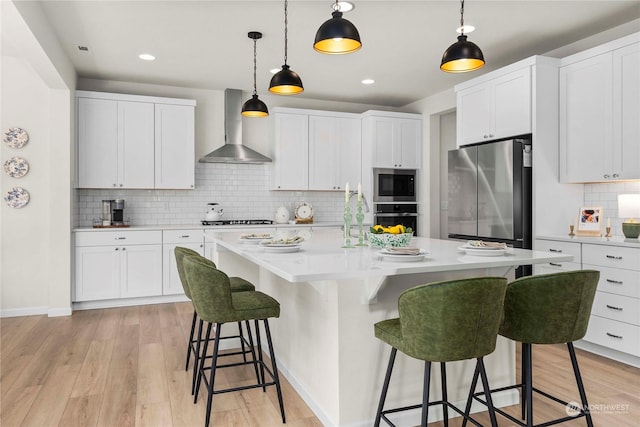 kitchen with wall chimney exhaust hood, stainless steel appliances, light hardwood / wood-style floors, decorative backsplash, and white cabinets