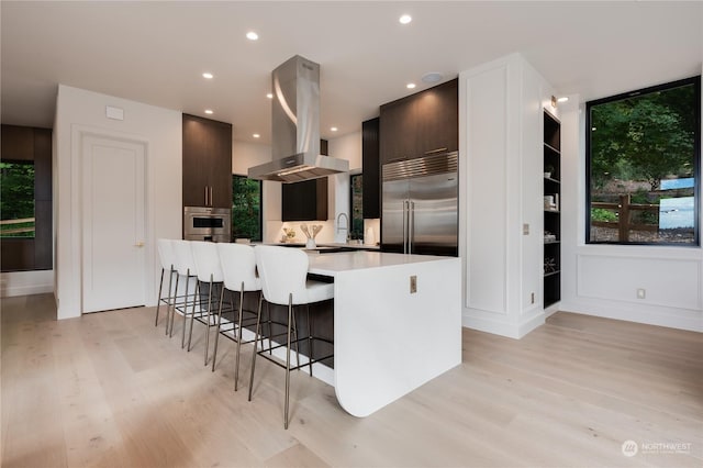 kitchen with appliances with stainless steel finishes, dark brown cabinets, island range hood, light hardwood / wood-style floors, and a kitchen island