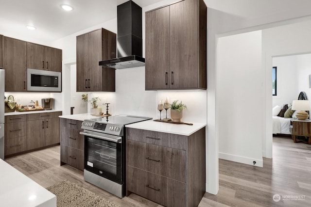 kitchen featuring wall chimney exhaust hood, dark brown cabinets, stainless steel appliances, and light hardwood / wood-style flooring