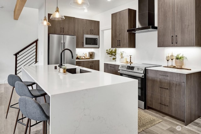 kitchen with wall chimney exhaust hood, hanging light fixtures, an island with sink, a breakfast bar area, and appliances with stainless steel finishes