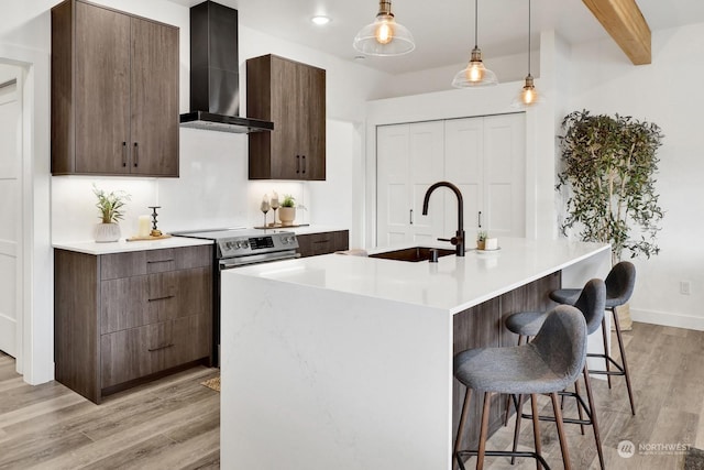 kitchen featuring stainless steel electric range oven, an island with sink, wall chimney range hood, and light wood-type flooring