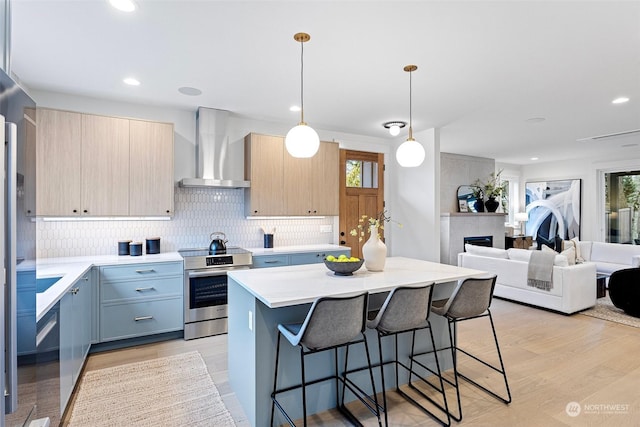 kitchen with wall chimney range hood, electric stove, light brown cabinetry, decorative light fixtures, and a kitchen island