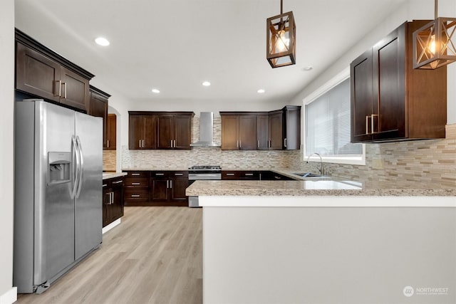 kitchen featuring kitchen peninsula, light wood-type flooring, stainless steel appliances, wall chimney range hood, and hanging light fixtures