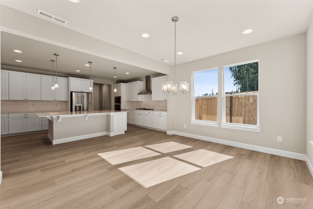 kitchen with wall chimney exhaust hood, hanging light fixtures, a large island with sink, light hardwood / wood-style floors, and appliances with stainless steel finishes