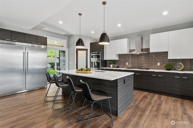 kitchen featuring dark brown cabinetry, stainless steel appliances, wall chimney range hood, an island with sink, and pendant lighting