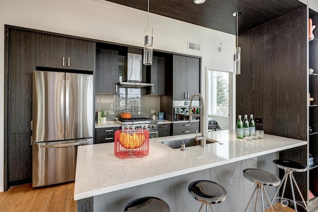 kitchen with sink, decorative light fixtures, light hardwood / wood-style flooring, wall chimney range hood, and appliances with stainless steel finishes