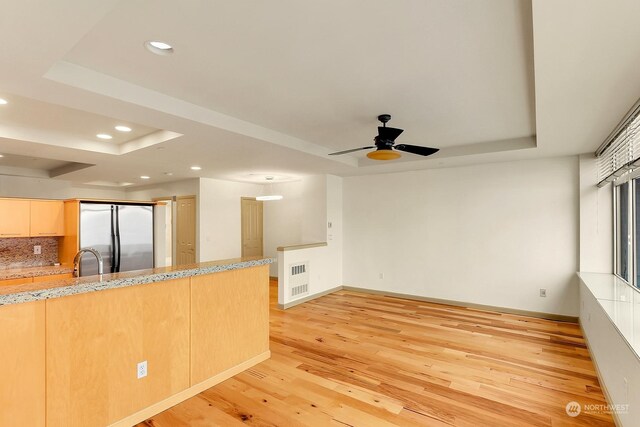 kitchen featuring a raised ceiling, ceiling fan, light hardwood / wood-style floors, light stone counters, and stainless steel refrigerator