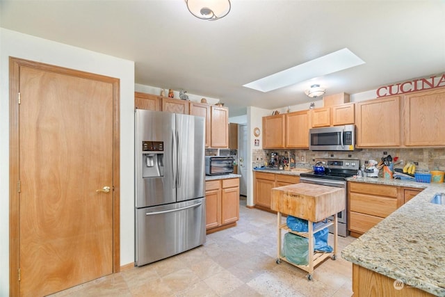 kitchen with light brown cabinetry, a skylight, appliances with stainless steel finishes, light stone countertops, and backsplash